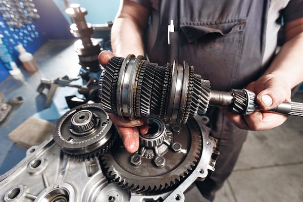 A person wearing overalls is holding a mechanical gear assembly with both hands. Various interconnected gears and mechanical parts are visible on a metal surface in a workshop setting.