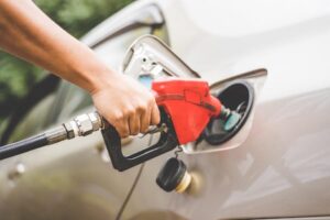 A close-up of a hand holding a red gasoline pump nozzle, filling the tank of a silver car. The car's gas cap is open, and the surrounding area of the vehicle is slightly visible. The background is blurred with greenery, possibly indicating an outdoor setting.