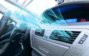 A car interior with the air conditioning system turned on. Blue graphical lines represent cool air flowing from the dashboard vents. The steering wheel, car keys, and dashboard controls are visible, with the key inserted in the ignition.