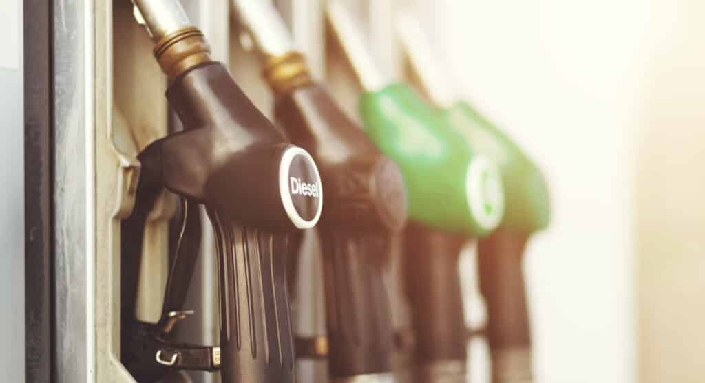 Close-up photo of fuel pump nozzles at a gas station, with one marked "Diesel" in focus. The nozzles are arranged in a row, featuring black and green handles. Sunlight filters through, creating a warm, soft ambiance.