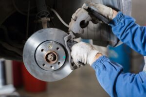 A mechanic wearing gloves and blue work clothes is installing a brake caliper onto a vehicle's brake rotor. The vehicle is elevated, and the focus is on the brake assembly components. The background is blurred, suggesting a workshop environment.