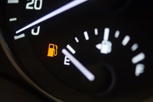 Close-up of a car dashboard fuel gauge showing near empty, with an illuminated yellow low fuel warning light. The gauge needle points towards the "E" (empty) mark.