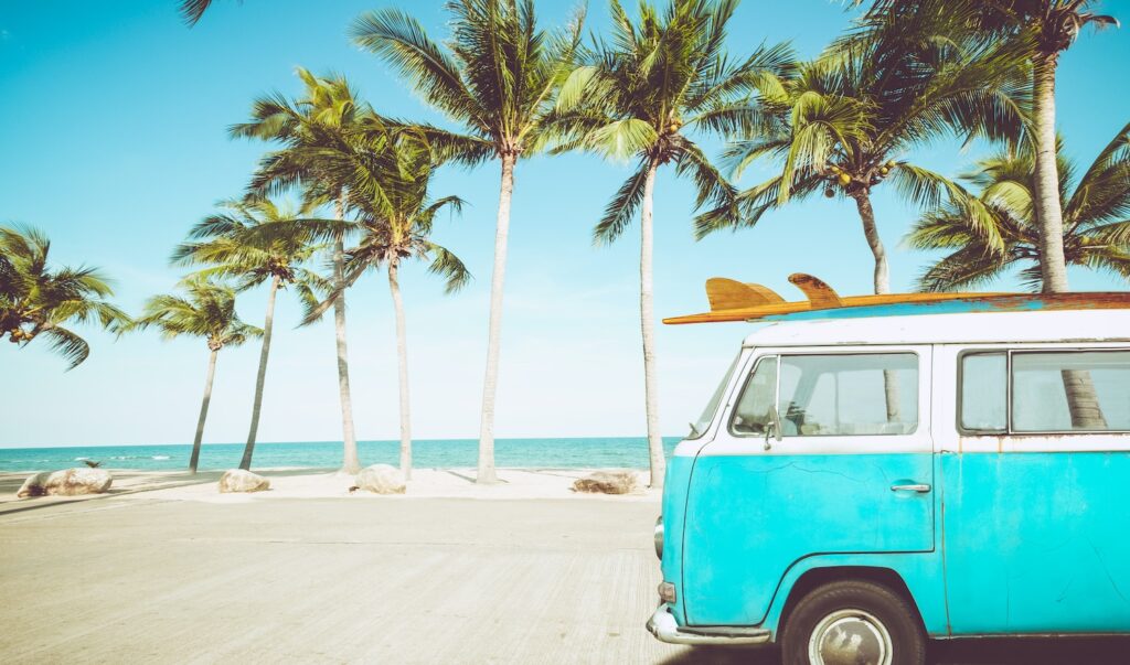 A bright blue vintage van with a surfboard on top is parked by a scenic beach lined with tall palm trees. The sky is clear and blue, and the ocean is visible in the background, creating a tropical, carefree atmosphere.