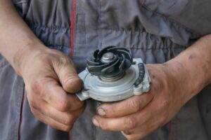 A person wearing a gray work uniform and with dirty hands is holding a mechanical part. The device has a fan-like component on top which appears to be part of an engine or similar machinery.