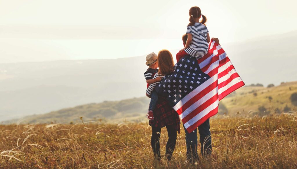 A family of four stands in a field, facing away from the camera. The father holds an American flag, while the mother carries a small child and another child sits on his shoulders. They all appear to be enjoying the scenic landscape in the distance.