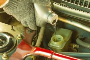 A person wearing a glove is pouring fluid from a grey bottle into a vehicle's engine compartment. The image focuses on the fluid being poured with car parts and components visible in the background.