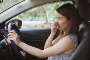 A woman is seated in the driver’s seat of a car, holding the steering wheel with her right hand while pinching her nose with her left hand, appearing bothered by an unpleasant smell. The car's window is up, and trees are visible outside.