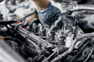 A gloved hand working on the engine of a vehicle. The image shows various components of the engine, including valves, springs, and tubes, with the person's hand adjusting a part. The setting is a mechanic workshop.