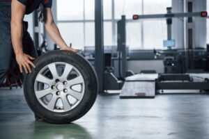 A person with cropped view rolling a car tire inside an automotive workshop. The workshop is equipped with various tools and machinery, and the background shows a vehicle lift and some workstations. The scene has a professional and industrial atmosphere.