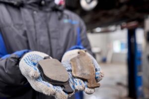 A person wearing a hooded jacket and white, blue-speckled gloves is holding two brake pads. The left brake pad appears new, while the right brake pad is worn and rusty. The background is blurred, featuring a car and a garage setting.