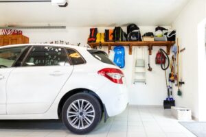 A white car is parked inside a tidy garage. The garage wall is neatly organized with various items, including a ladder, leaf blower, and various bags and storage boxes placed on shelves. The floor is clean and the garage is well-lit.