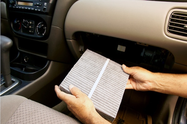 A person is holding a cabin air filter, which has been removed from the glove compartment of a car. The car's dashboard, control buttons, and partially open glove compartment are visible in the image.