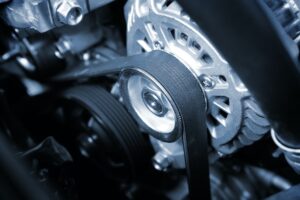 Close-up of a car engine timing belt system. The image shows various components including pulleys and the belt, which appears in clear focus, highlighting the gears and tensioners in a detailed metallic, monochromatic blue hue.