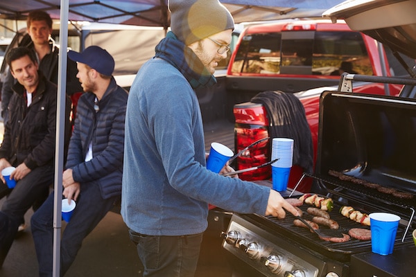 A man in a beanie and glasses cooks food on a grill at the back of a red truck, holding a blue cup in one hand and a spatula in the other. Three men in jackets and blue cups sit and chat nearby, with tents and other tailgating supplies visible in the background.