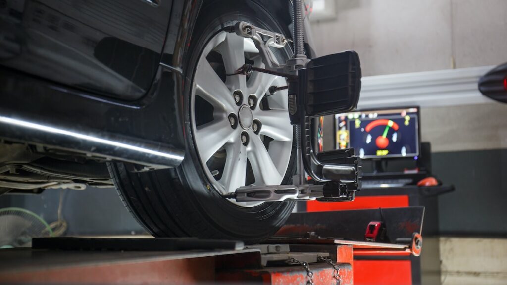 A car is elevated on a lift, undergoing a wheel alignment. A computer screen with diagnostic data is visible in the background. The alignment tool is attached to the car's wheel, which is highlighted in focus. The setting appears to be an automotive workshop.