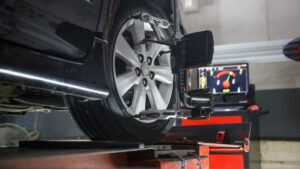 A car is elevated on a lift, undergoing a wheel alignment. A computer screen with diagnostic data is visible in the background. The alignment tool is attached to the car's wheel, which is highlighted in focus. The setting appears to be an automotive workshop.