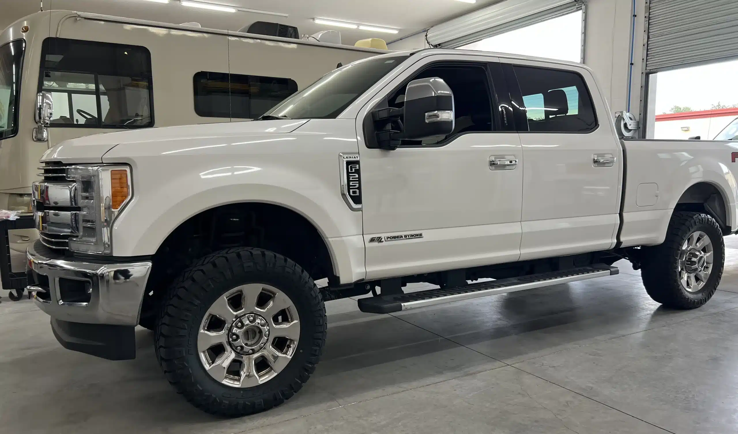 A white pickup truck with large off-road tires is parked indoors, near a camper van. The truck has chrome accents and side steps, and its interior is visible through tinted windows.