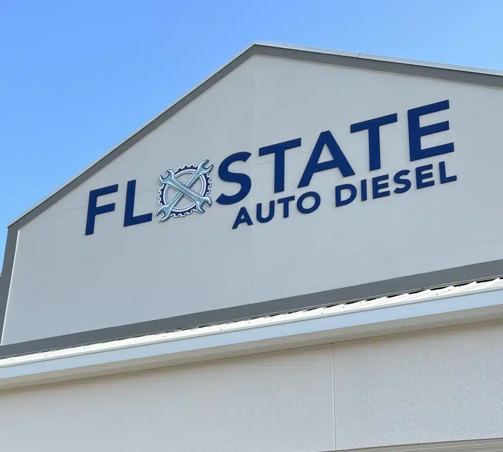 A building facade with a gable roof displaying a sign that reads "FLOSTATE AUTO DIESEL" in dark blue letters. The "O" in "FLOSTATE" features a cog and wrench logo. The building is light-colored, and the sky in the background is clear and blue.