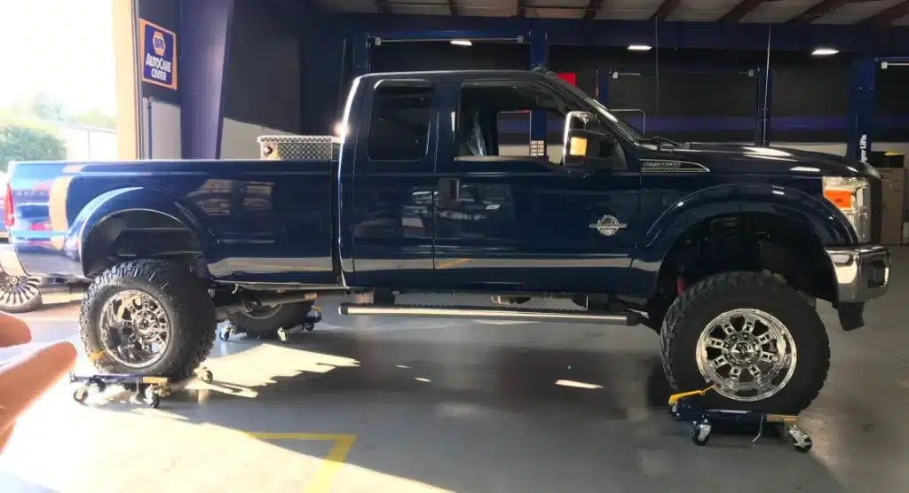 A dark blue lifted pickup truck is parked inside a garage with bright overhead lighting. The truck has large, rugged tires and shiny chrome rims. It is elevated on wheeled platforms, possibly for maintenance or detailing.