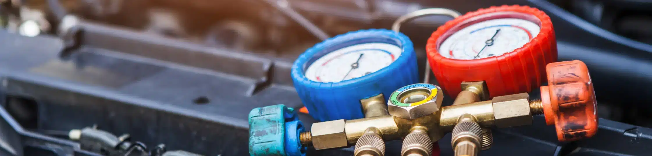 Close-up of HVAC manifold gauges with a red and blue dial, used for measuring pressure in air conditioning systems. The background shows part of an engine, slightly out of focus.