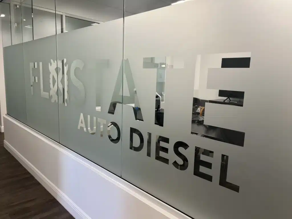 Frosted glass office partition with "FloState Auto Diesel Repair" cut out, offering a peek into a modern workspace with desks and chairs. The dark wooden flooring complements the sleek vibe, making it an ideal spot for anyone searching for top-notch auto repair in St. Cloud.