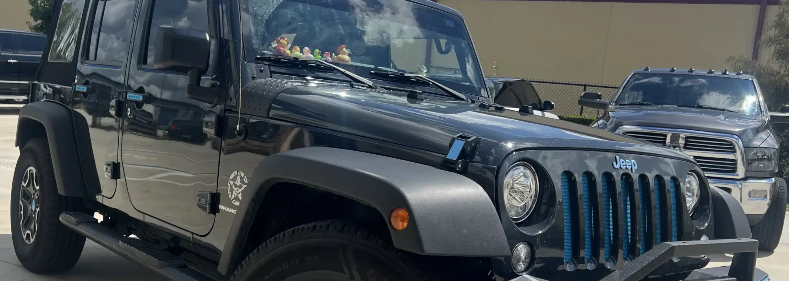 Image of a Jeep Wrangler, emphasizing its distinct round headlights and grille, is parked next to another vehicle. The backdrop subtly hints at an auto and diesel repair setting, with the edge of another car partially visible in the background.