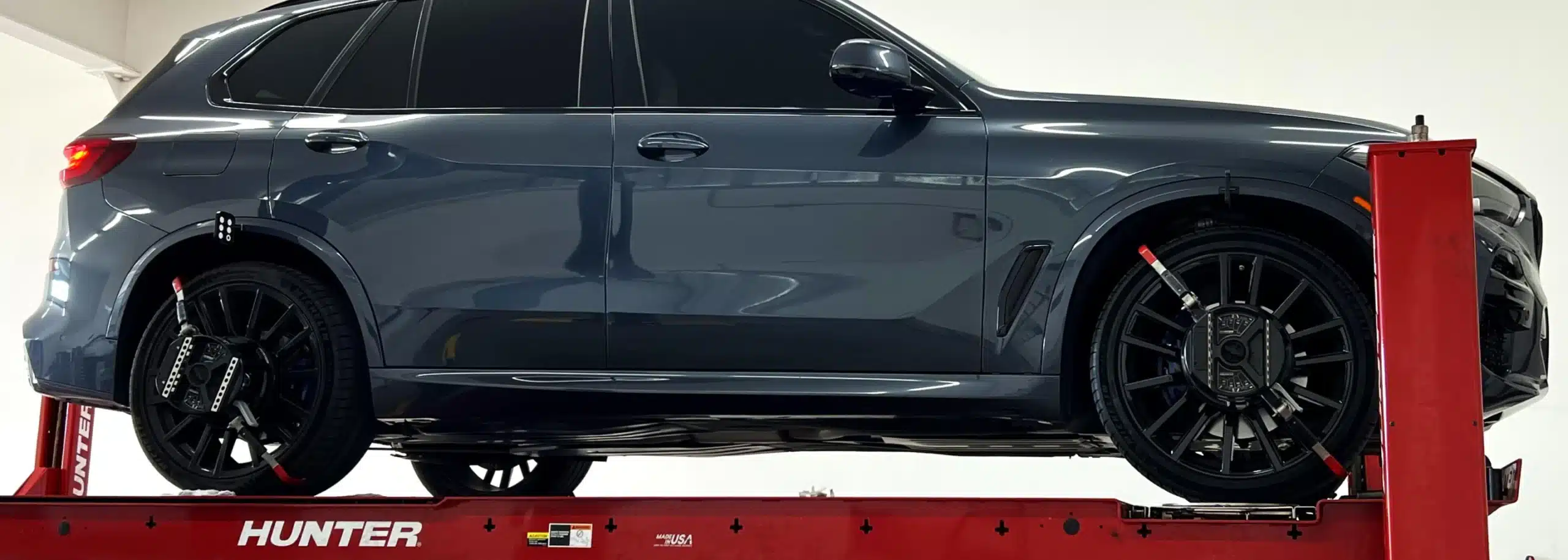 A sleek car on a hydraulic lift in an automotive service center for wheel alignment. The vehicle is positioned sideways, showcasing its side profile with dark-tinted windows. The lift, marked HUNTER, supports precision work.