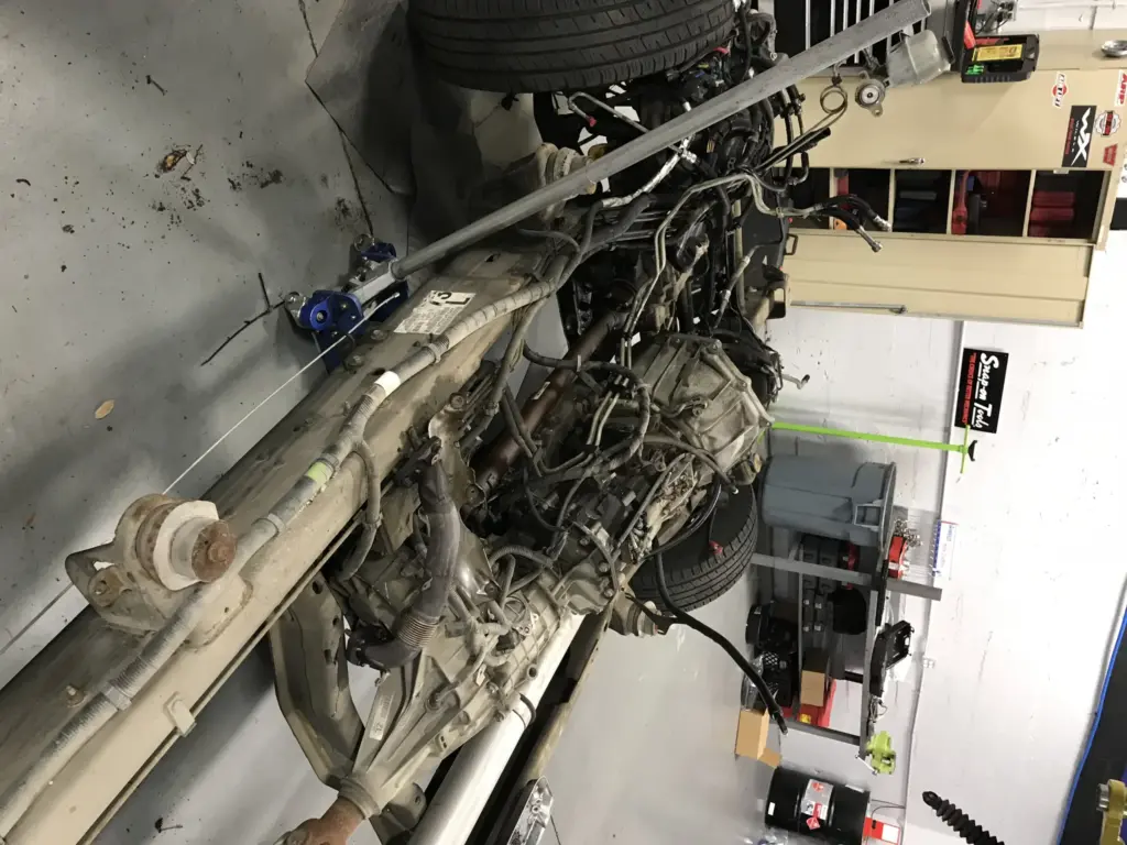 Image of a vehicle chassis and engine with various components exposed in a mechanic's workshop. Tools and equipment are scattered around, with shelves and cabinets visible in the background. The vehicle appears to be undergoing repair or maintenance work.