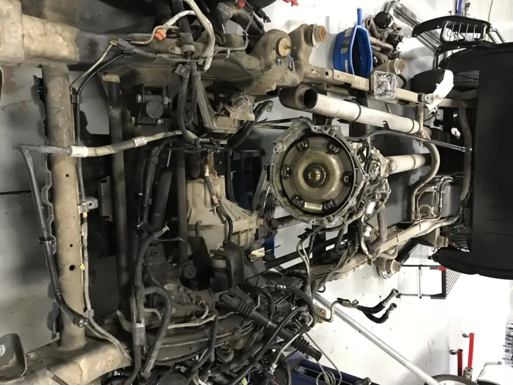 An image of a vehicle's underside on a lift in an auto repair shop. The exposed drivetrain components, wires, and parts are visible. Mechanical tools and equipment are seen around the garage floor, and various car parts are scattered nearby.