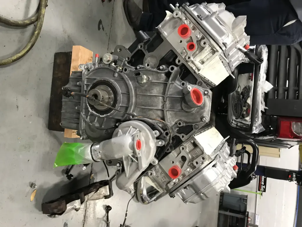 A partially assembled car engine on a workshop floor. The engine features various components and metallic parts, some tagged with labels, and several openings are covered with red caps. Two technicians' lower legs are visible in the background.