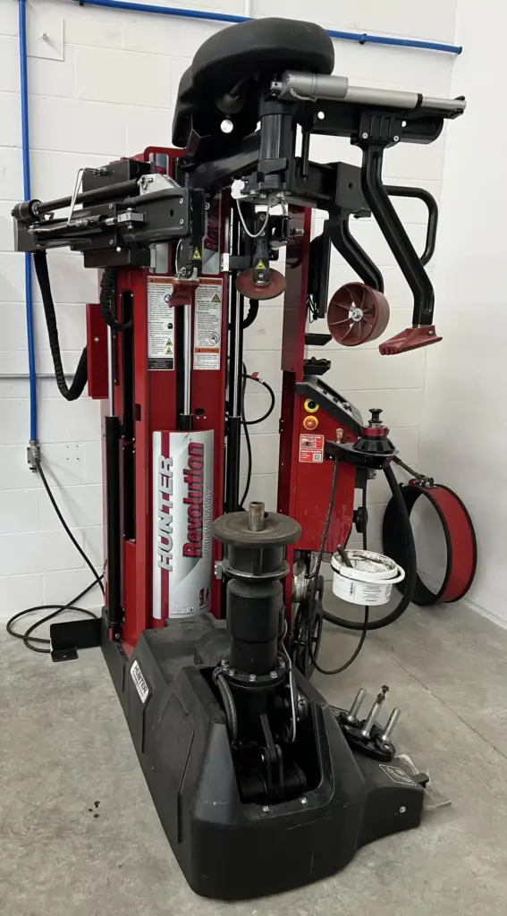 A red and black tire changing machine stands on a concrete floor next to a white wall. The machine features various levers, clamps, and a central vertical column with the word "Revolution" and brand name "Hunter" visible. Various tools and attachments are nearby.