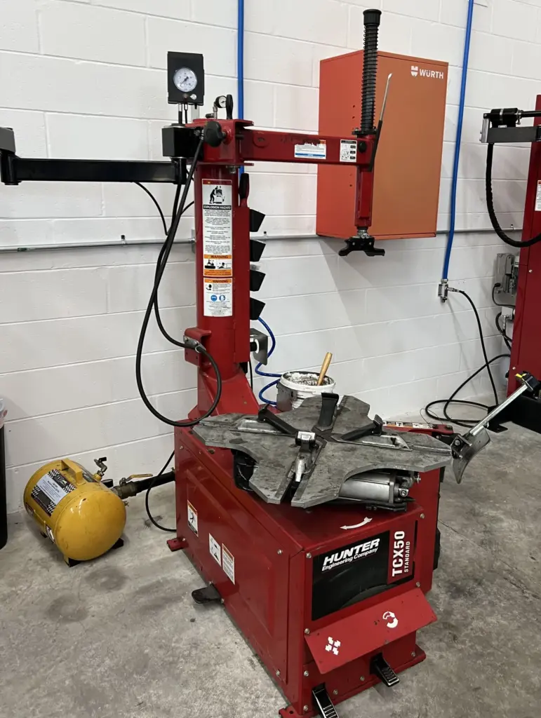 A red Hunter tire changer machine in a garage with various tools and equipment around it. There's a gauge at the top, assorted levers and controls, and a tire rim clamped in place. A red cabinet and an air compressor tank are in the background.