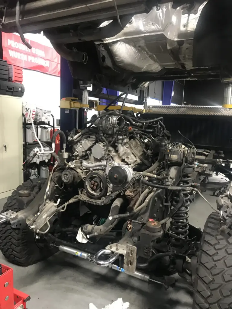 A vehicle on a lift with its front end disassembled, exposing the engine and various mechanical components. The shop is equipped with tools and machinery, and a red banner is partially visible in the background.