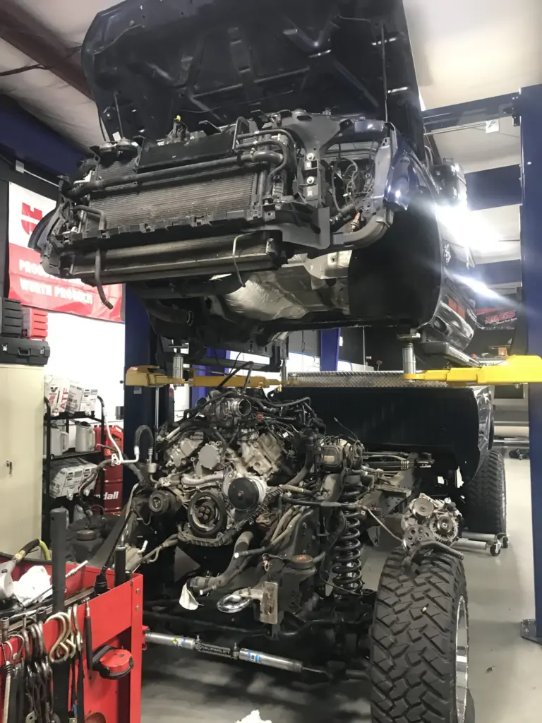 A vehicle is raised on a hydraulic lift in a garage, exposing the undercarriage and engine bay. The front bumper and grill have been removed, and the engine is partly disassembled. Tools and equipment are visible around the work area.