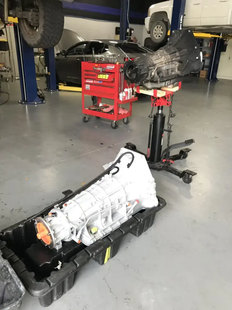 An automotive repair shop with a transmission on a hydraulic lift in the foreground. Another transmission rests in a large container on the floor. Various tools and equipment, including a red tool chest and a car on a lift, are visible in the background.
