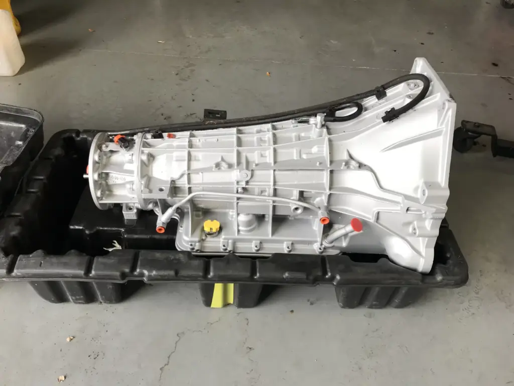 A car transmission with a white and gray exterior, black wiring, and red caps, lies on a black plastic tray in a garage setting. The transmission appears clean and well-maintained.