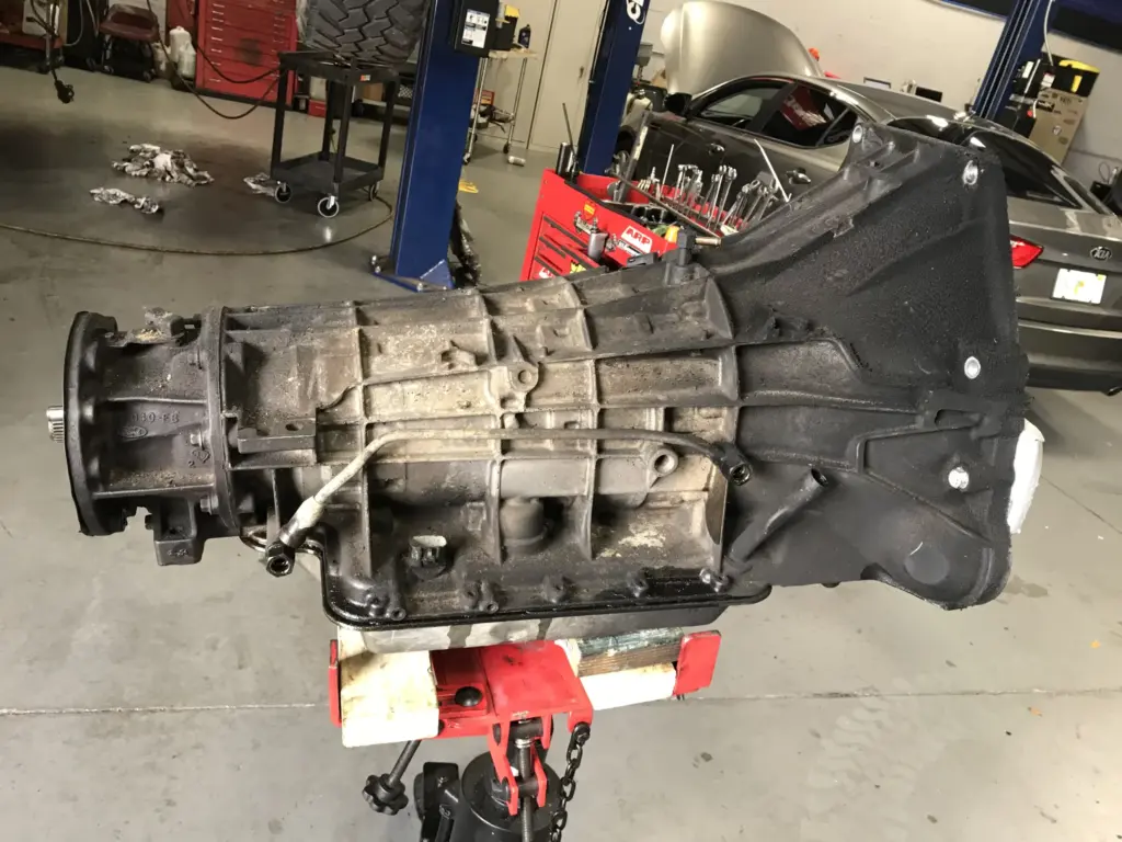A car transmission sits on a red hydraulic lift inside an automotive repair shop. The transmission appears to be greasy and used. In the background, vehicle lifts, tool cabinets, and partially visible cars can be seen. The shop floor is clean and organized.