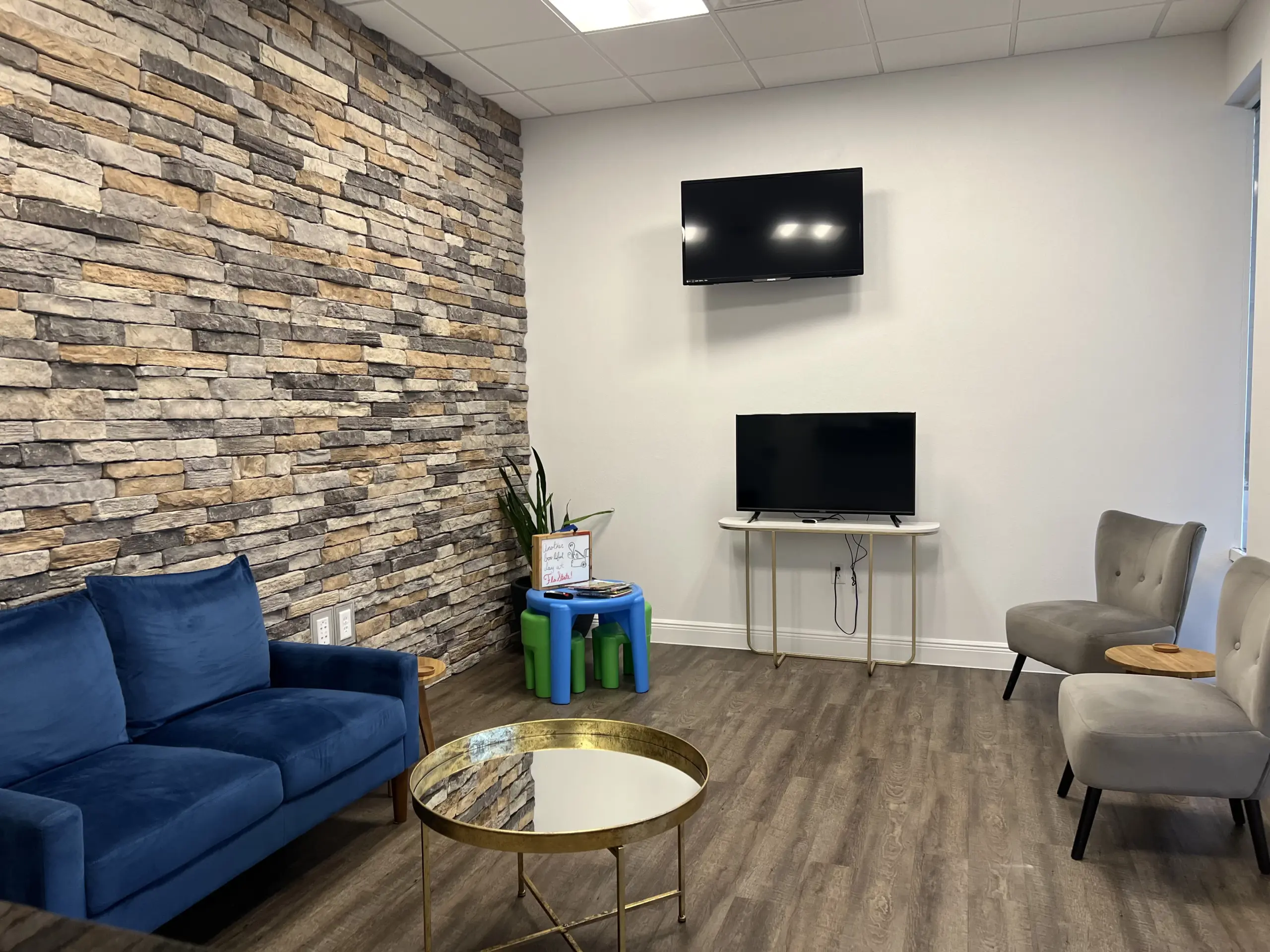 A modern waiting room for an auto repair near me features a stone accent wall, blue sofa, gray chairs, round gold coffee table, and a small TV on a stand. A children's play area with colorful stools enhances the space, while a larger TV is mounted on the wood-style floor wall.