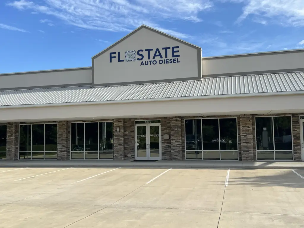 A storefront for "FloState Auto Diesel" under a blue sky. The building has a brick facade, large glass windows, and an angled metal roof. The pavement in front has several empty parking spaces.