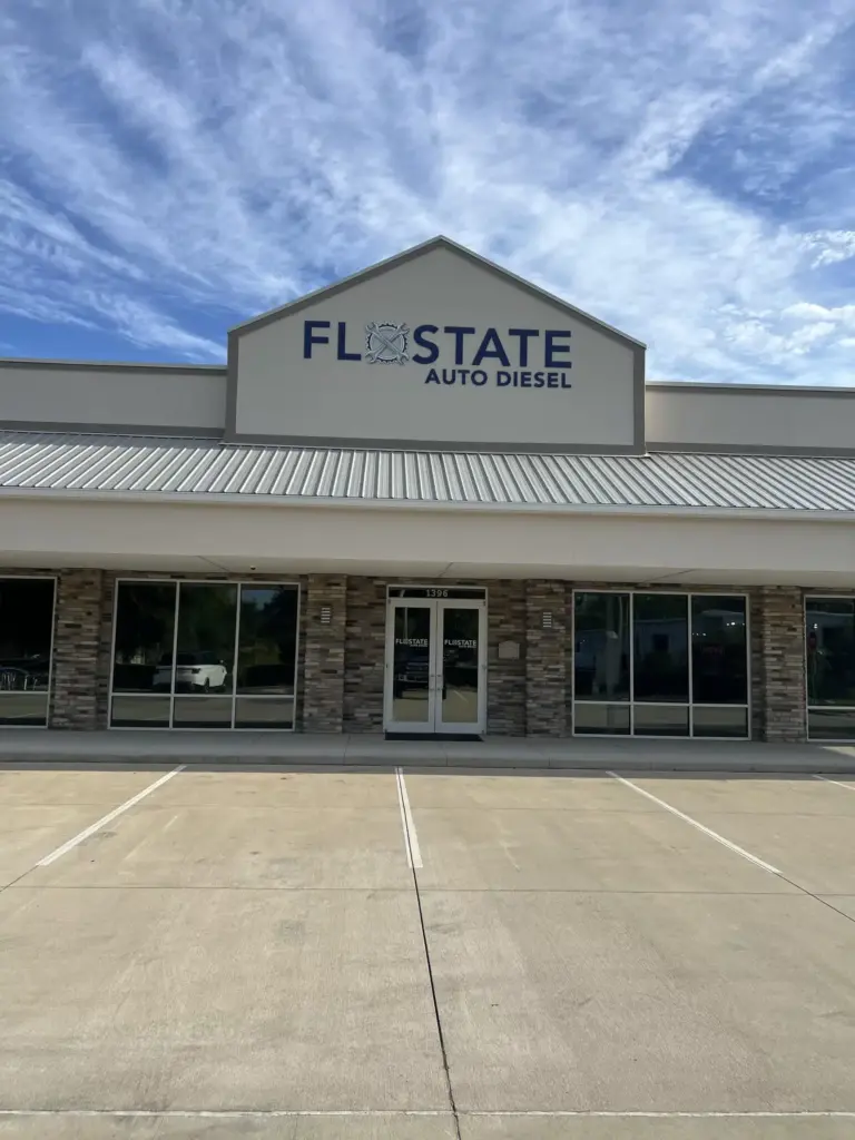A storefront with a sign that reads "FlowState Auto Diesel" above the entrance. The building features a stone façade, large windows, and a wide concrete parking area in front. The sky above is partly cloudy.