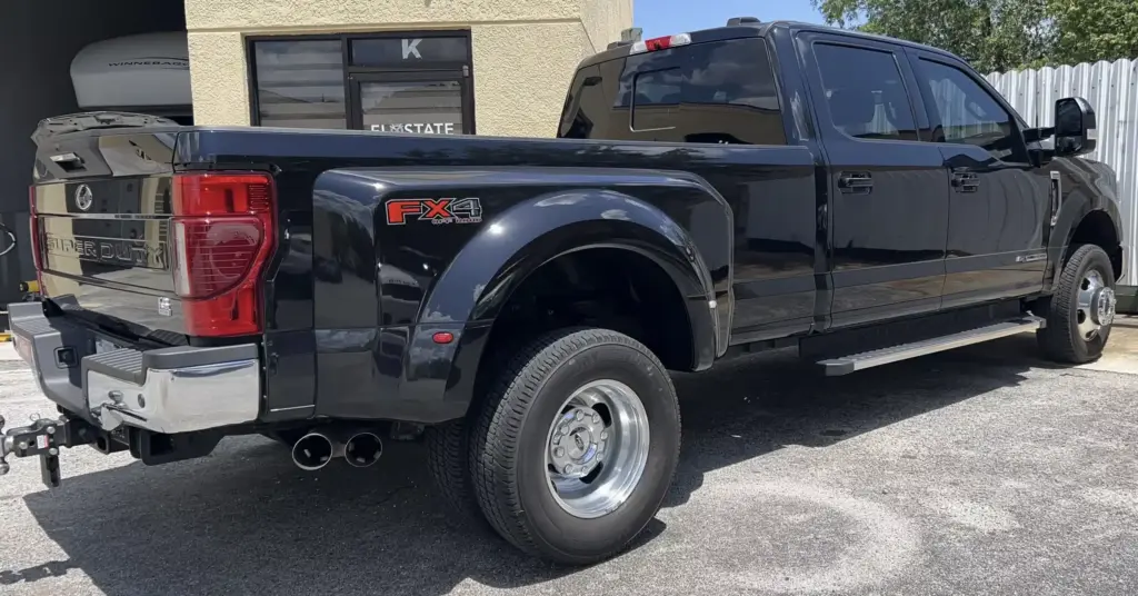 A black Ford Super Duty FX4 pickup truck is parked outside a beige building. The rear of the truck is visible, showcasing its dual rear wheels, large tailgate, exhaust pipes, and tow hitch.