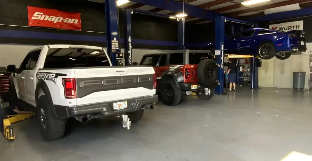 A garage with three vehicles: a white Ford Raptor, a red Jeep, and a blue car on a lift. The space has clear, organized flooring with vehicle maintenance equipment. Snap-on and Wurth banners are visible on the walls.