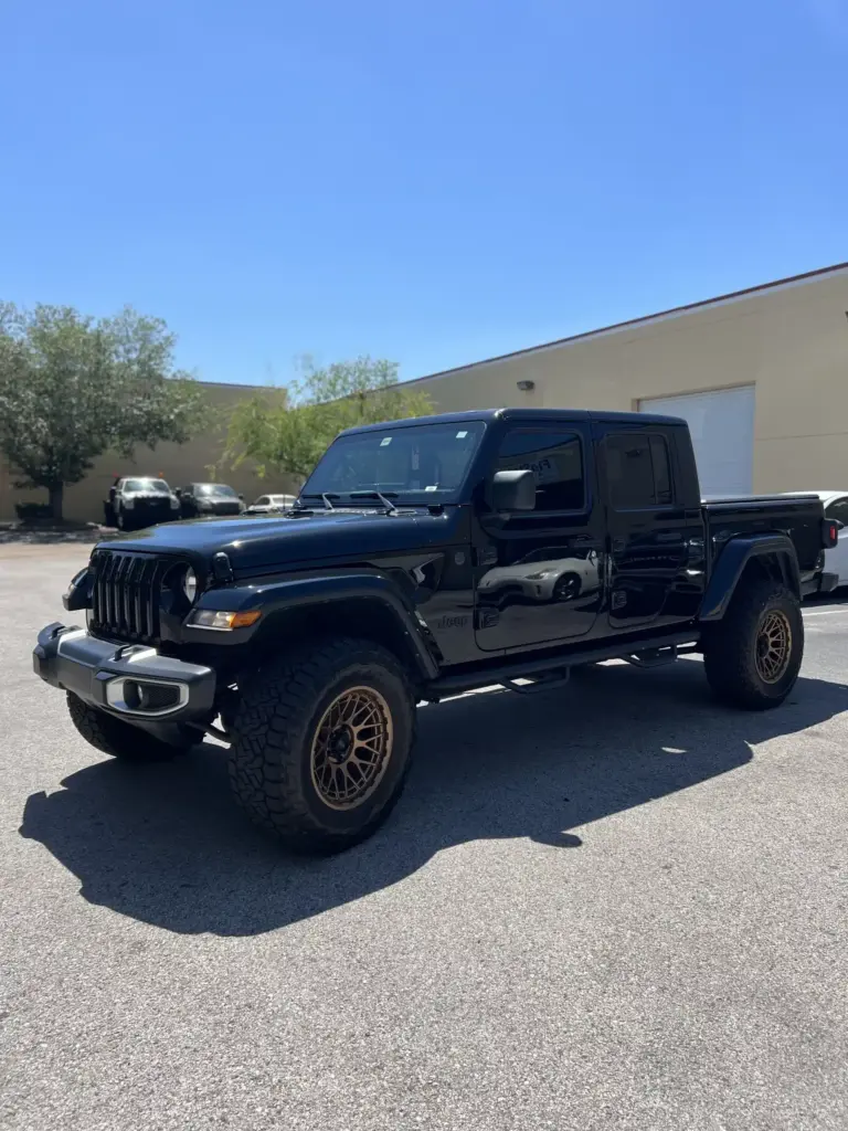 A black Jeep Gladiator parked in a sunny outdoor lot. The truck has large, rugged tires with gold rims. In the background, shrubs and part of a beige building with white garage doors are visible.