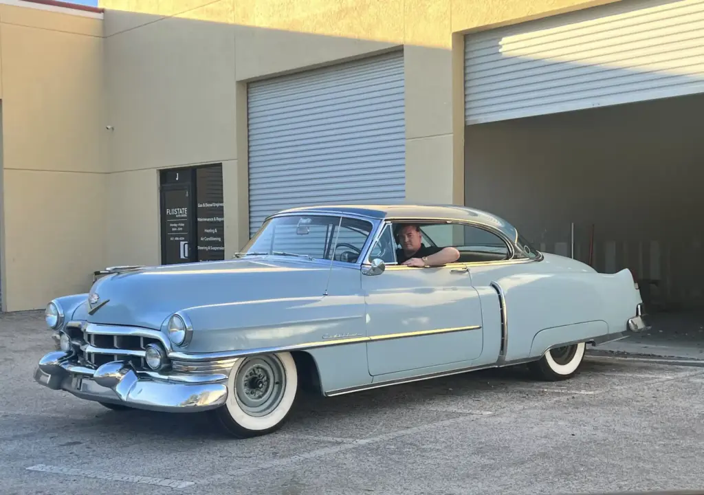 A man drives a classic light blue Cadillac with a shiny chrome exterior and whitewall tires. The car is parked near a beige building with a closed garage door. The driver is seen through the open car window.