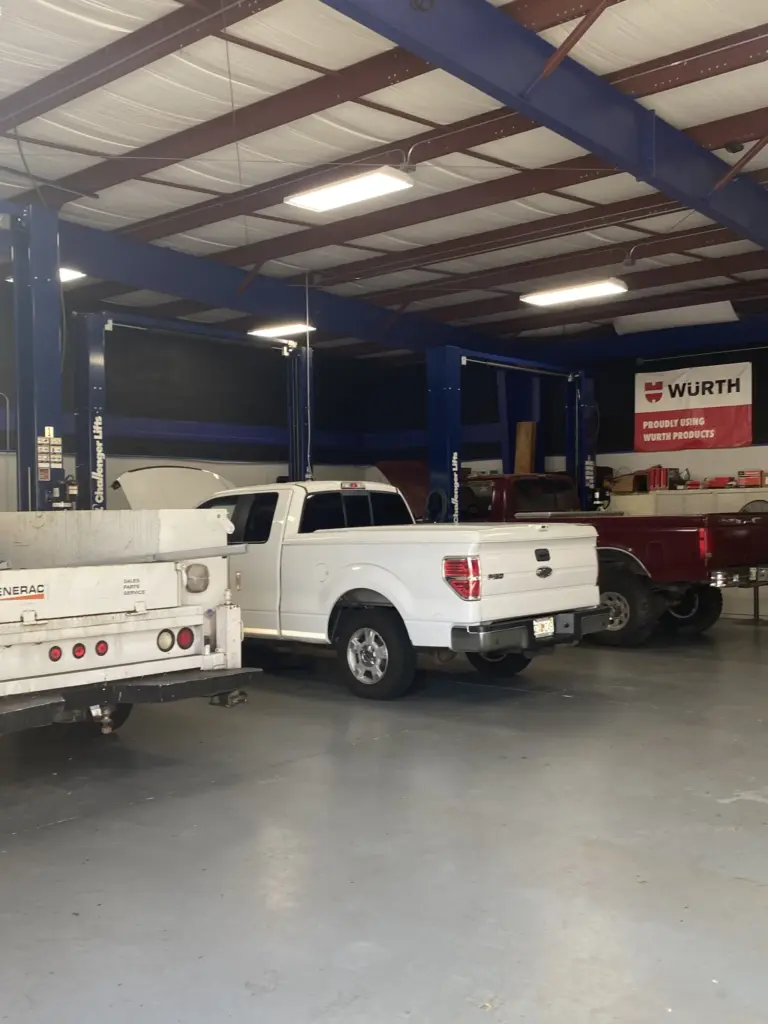 An auto repair shop with a high ceiling and exposed beams. Three trucks are being serviced: a white one in the center, a red one to the right, and a white flatbed truck to the left. The shop has a banner that reads "Wurth" and various tools and equipment are visible.