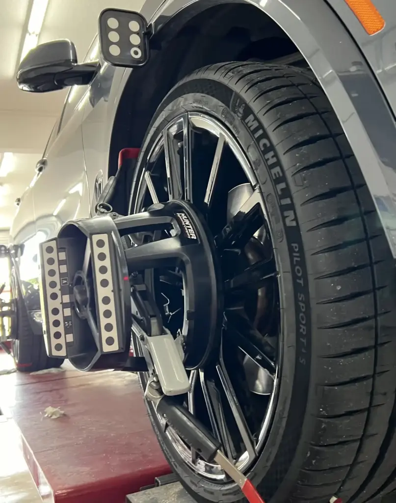 Close-up of a car wheel equipped with a Michelin Pilot Sport tire, mounted on a vehicle undergoing alignment in an auto shop. The tire is attached to alignment measurement equipment, with part of the car's body and alignment platform visible in the background.