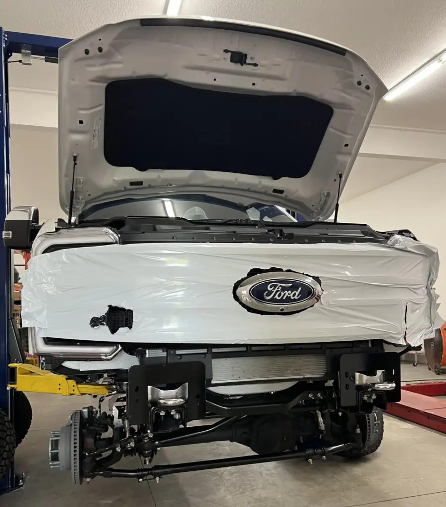 A white Ford vehicle with its hood open is being repaired in a garage. The front bumper and several components are missing, exposing the internal mechanical parts. The car is elevated on a lift, and tools are visible in the background.