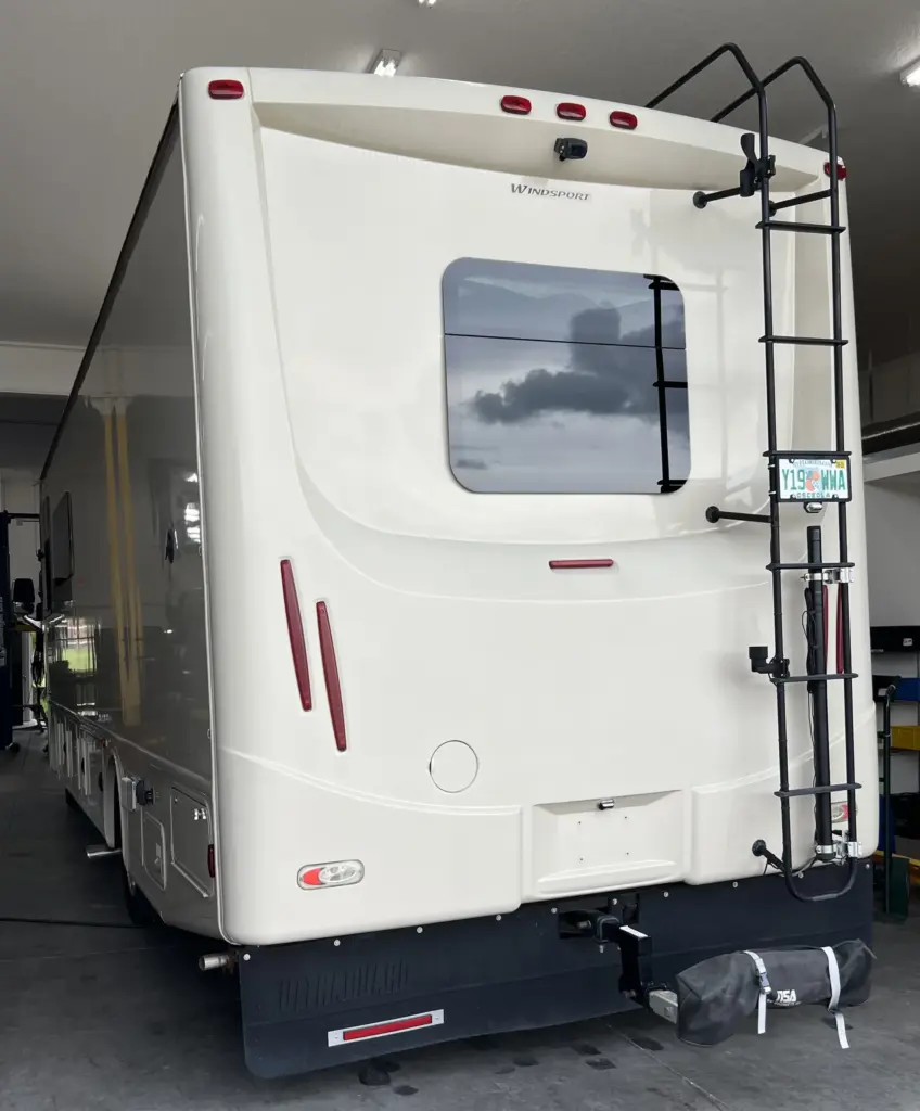 A large, beige RV is parked inside a garage. The rear of the RV features a window, a ladder on the right side, taillights, and a Florida license plate. The RV has a spare tire cover attached to the back bumper. The interior garage lighting is visible.
