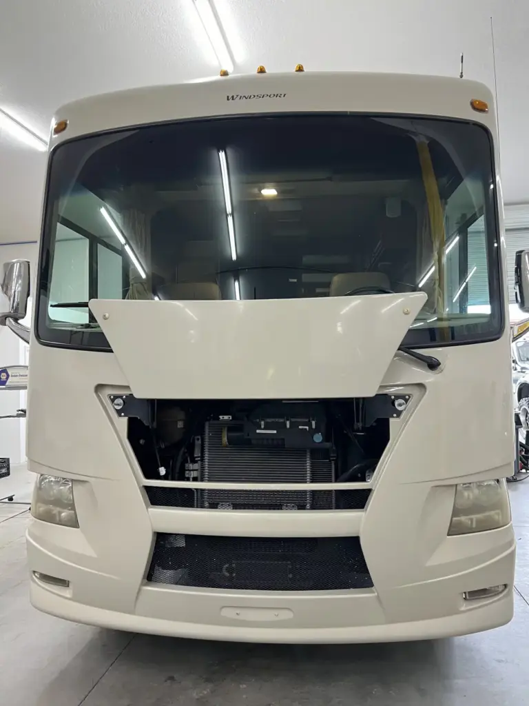 Front view of a motorhome with the hood open, revealing the engine compartment. The vehicle is inside a garage or workshop, with tools and equipment visible in the background. Bright, even lighting illuminates the space.